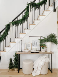 Modern farmhouse entryway with black metal staircase railing draped in evergreen garland, wooden treads, neutral console table styled with candles, framed art, evergreen vase and cozy white throw
