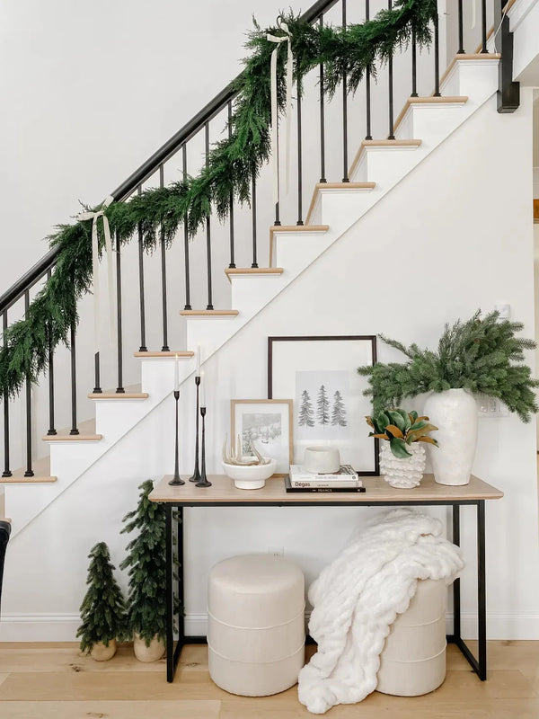 Modern farmhouse entryway with black metal staircase railing draped in evergreen garland, wooden treads, neutral console table styled with candles, framed art, evergreen vase and cozy white throw