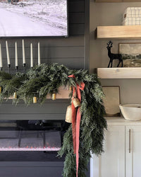 Festive Christmas mantel with lush cedar garland, red ribbon and bells draped over a modern black shiplap electric fireplace with floating wood shelves and decor