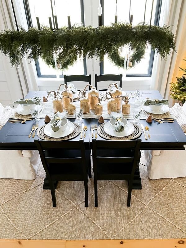 Modern farmhouse Christmas dining tablescape with hanging greenery chandelier, wooden bell centerpiece, woven chargers, white dinnerware, and gold flatware on a black table