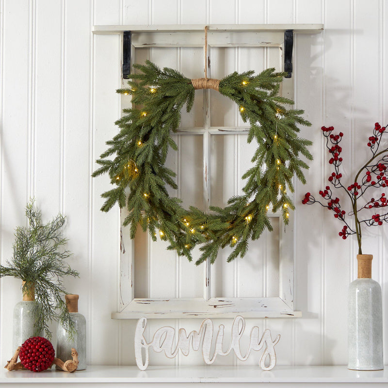 Lit pine Christmas wreath with warm LED lights hanging on distressed white window frame over mantel with "family" sign and berry decor