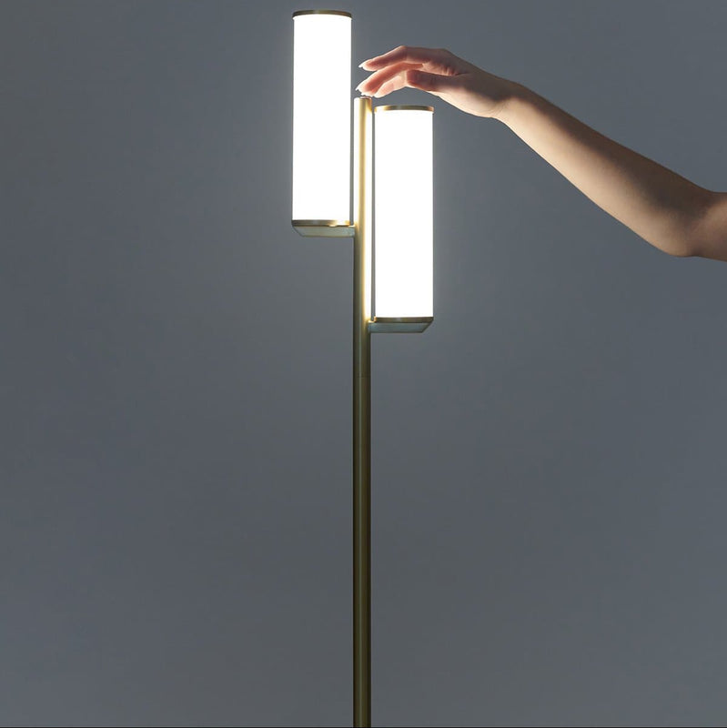 Modern brass floor lamp with dual cylindrical LED shades, hand adjusting dimmable ambient lighting for minimalist living room decor.