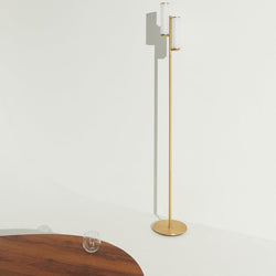 Minimalist interior featuring a modern brass floor lamp casting a long shadow on a white wall, with a wooden coffee table and glass tumbler, contemporary home decor