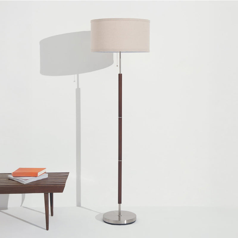 Modern floor lamp with beige drum shade, dark wood pole and brushed metal base next to mid-century wooden bench with books.
