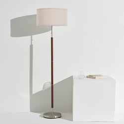 Minimalist modern floor lamp with beige drum shade, wooden stem and brushed metal base beside white pedestal with books and glass, textured gray rug, contemporary living room decor