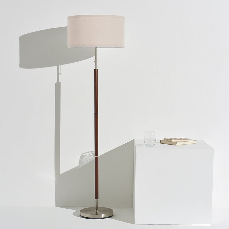 Minimalist modern floor lamp with beige drum shade, wooden stem and brushed metal base beside white pedestal with books and glass, textured gray rug, contemporary living room decor