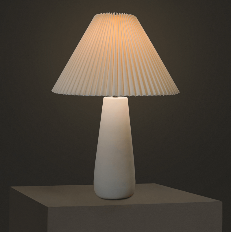 Minimalist white ceramic table lamp with pleated beige fabric shade on pedestal, warm ambient lighting for modern bedroom or living room decor
