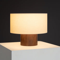 Mid-century modern table lamp with walnut cylindrical base and beige drum fabric shade casting warm ambient light on a white pedestal