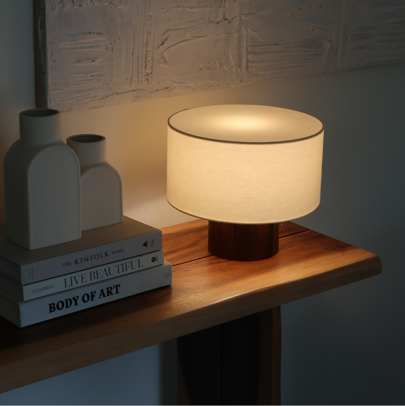 Minimalist wooden-base table lamp with linen drum shade casting warm ambient light on a wood console table next to stacked design books and ceramic vases, textured wall art behind