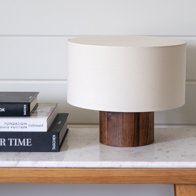 Minimalist modern table lamp with cylindrical dark wood base and white drum shade on a marble-top console table next to stacked coffee-table books, stylish home decor