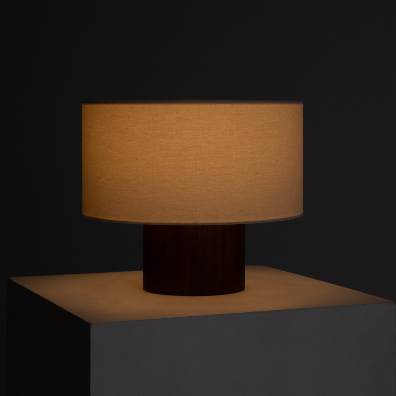 Minimalist modern table lamp with fabric drum shade and wooden base casting warm ambient light on white pedestal against dark background