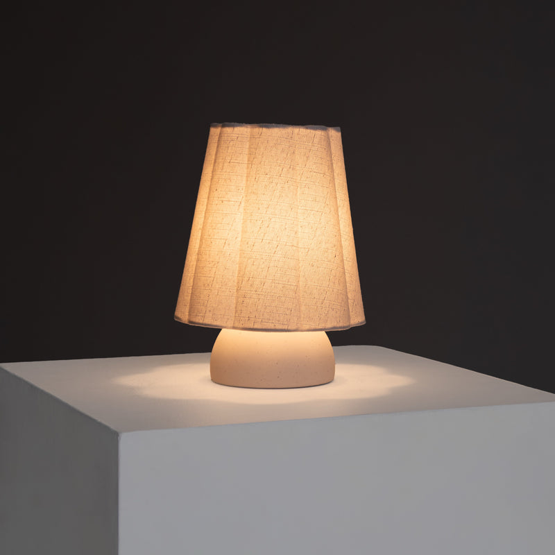 Minimalist beige fabric-shade table lamp with ceramic base on white pedestal, warm ambient bedroom and living room lighting
