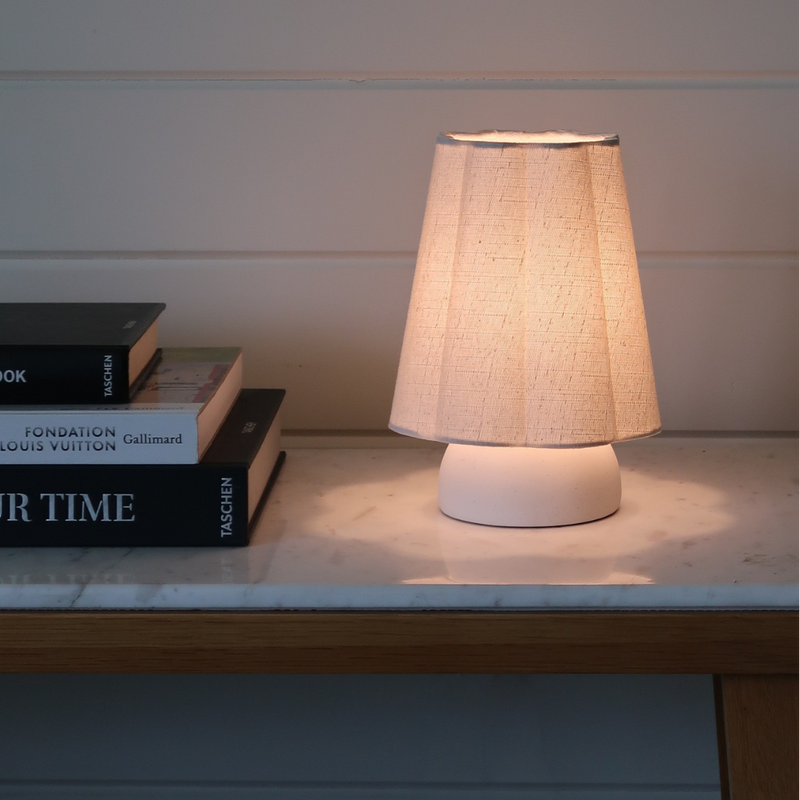 Minimalist beige fabric table lamp casting warm ambient light on a marble console table beside stacked art books against a white shiplap wall