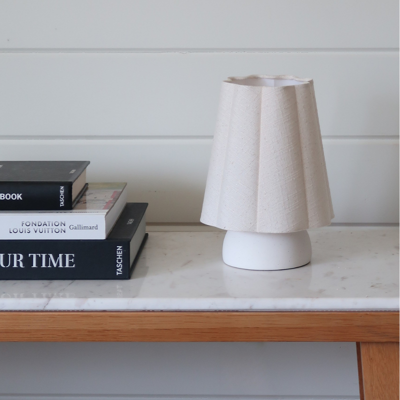 Minimalist white ceramic table lamp with pleated fabric shade on a marble-top wooden console beside stacked designer coffee table books — neutral modern home decor