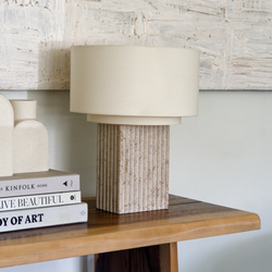 Beige linen drum table lamp with ribbed stone base on wooden console table beside stacked design books and textured abstract wall art