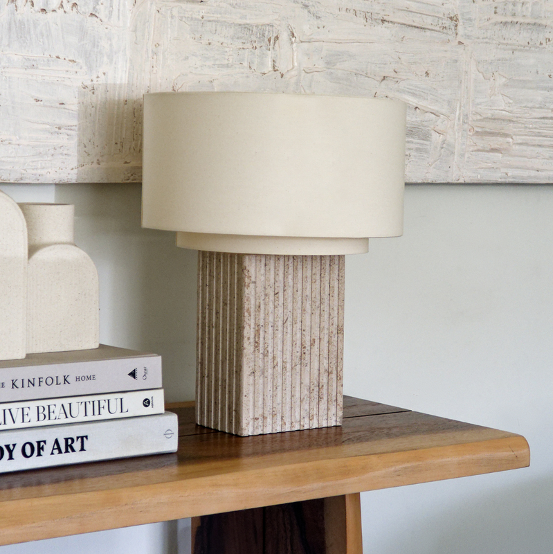 Beige linen drum table lamp with ribbed stone base on wooden console table beside stacked design books and textured abstract wall art