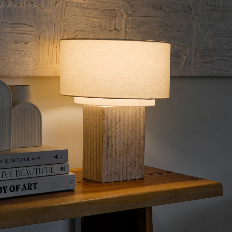 Modern minimalist table lamp with ribbed stone base and linen drum shade casting warm ambient light on a wooden console table beside stacked design books