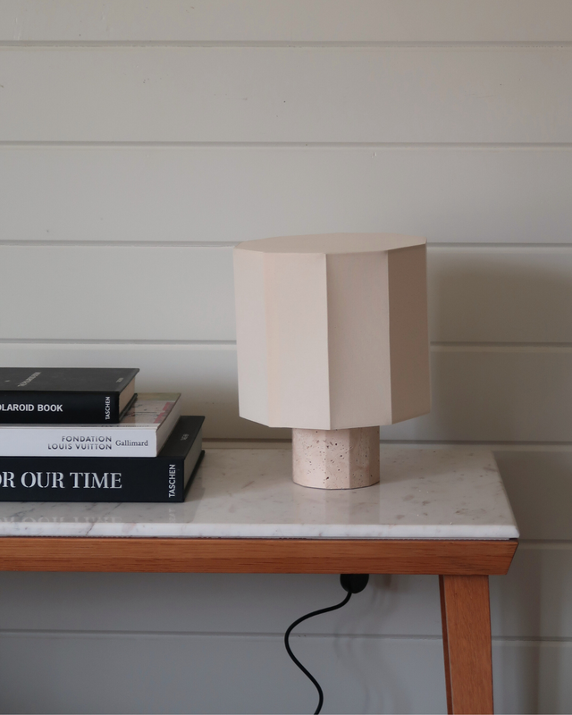 Minimal beige geometric table lamp on marble-top wooden console table beside stacked coffee table books against white shiplap wall, modern home decor interior styling