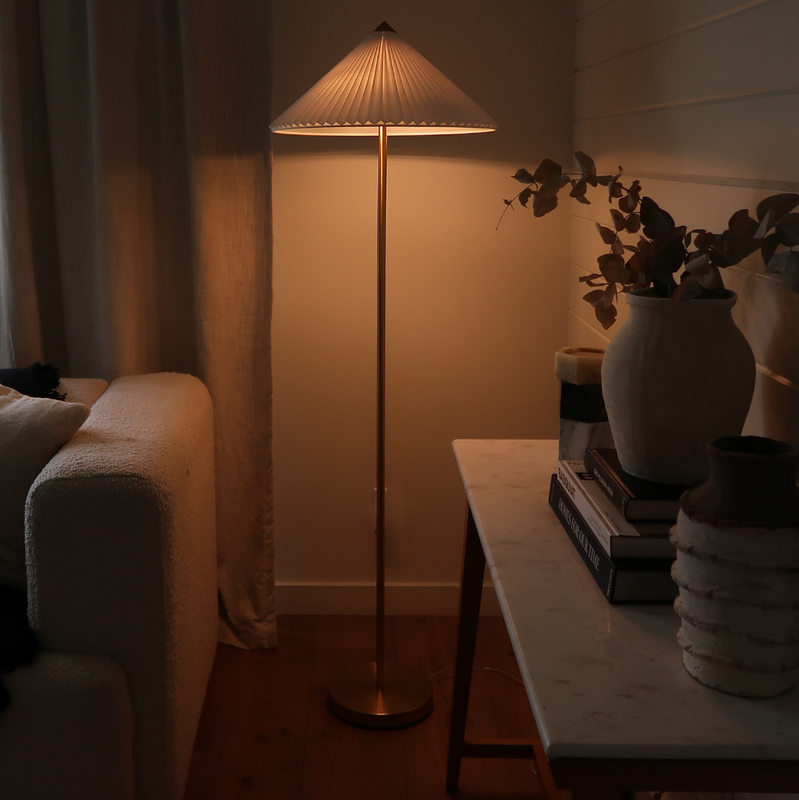Modern brass floor lamp with pleated shade casting warm ambient light beside sofa and console with ceramic vase in a cozy neutral living room