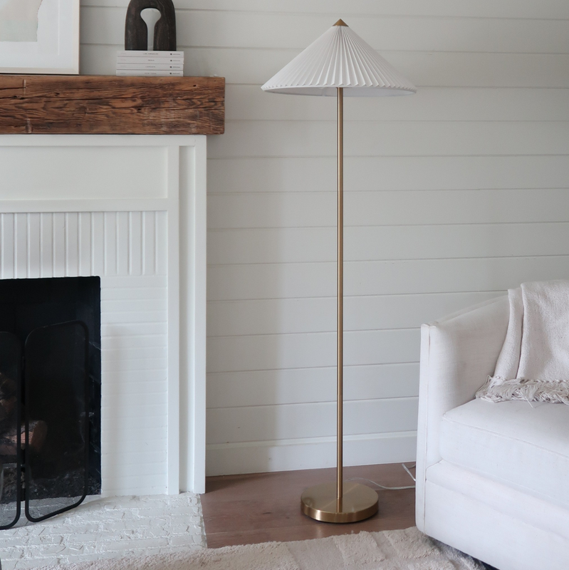 Modern brass floor lamp with pleated white shade next to a cream sofa and rustic wooden mantel above a white fireplace in a minimalist living room