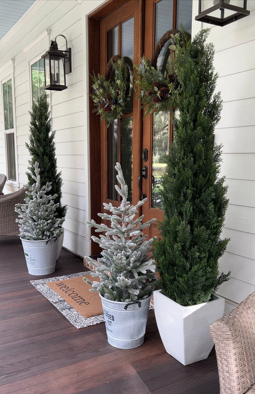 Farmhouse front porch with a wooden double door and evergreen wreaths, potted columnar topiaries and flocked mini Christmas trees in metal buckets on a welcome mat under black lantern sconces and white siding