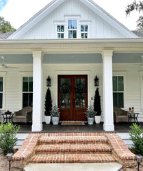 Modern farmhouse front porch entry with brick steps, white columns, dark wood double doors with wreaths, potted evergreens and wicker seating