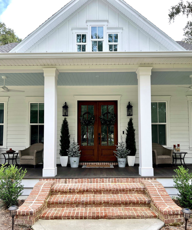 Modern farmhouse front porch entry with brick steps, white columns, dark wood double doors with wreaths, potted evergreens and wicker seating