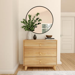 Minimalist entryway with round wall mirror above a light wood three-drawer dresser, decorative vase with eucalyptus branches, small black vase and bowl on stacked books, neutral walls and hardwood floor, modern home decor