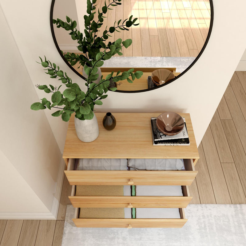 Overhead view of a light-wood three-drawer dresser with open drawers revealing folded linens, round wall mirror, decorative vase with green eucalyptus and stacked books on top in a modern minimalist entryway with hardwood floors