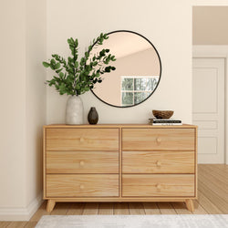 Modern minimalist bedroom with wooden dresser, round mirror, and decorative plants.