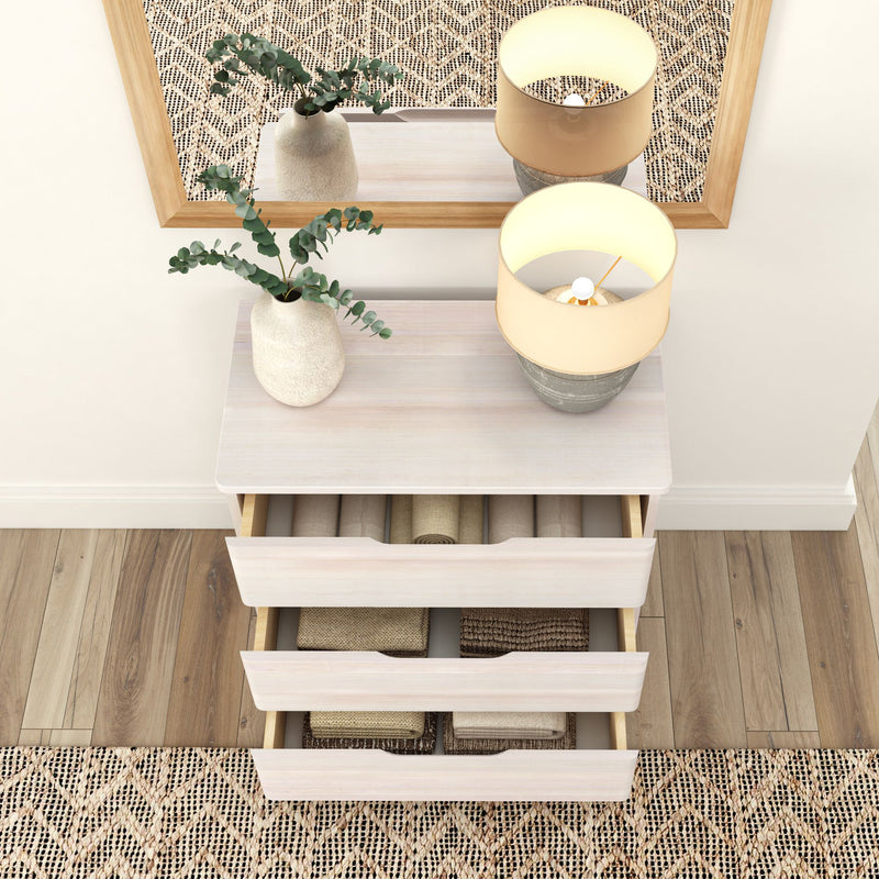 Modern dresser with open drawers displaying neatly folded towels, a vase with greenery, and a stylish lamp, set on wooden flooring and patterned rug.