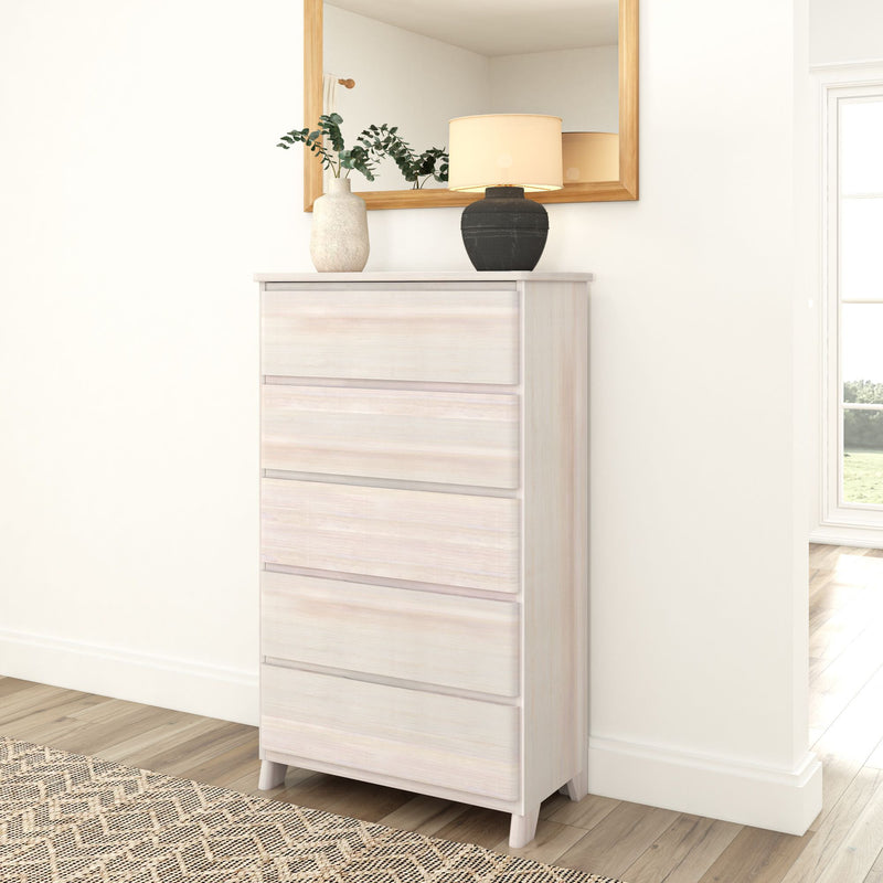 White wooden dresser with five drawers, decorated with a black lamp and ceramic vase, placed against a white wall beside a large window in a modern bedroom.