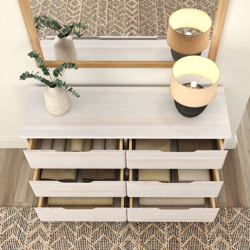 Modern white dresser with six open drawers containing neatly folded beige and brown textiles, decorated with a small plant and lamp, reflecting in a large mirror on a wooden floor.