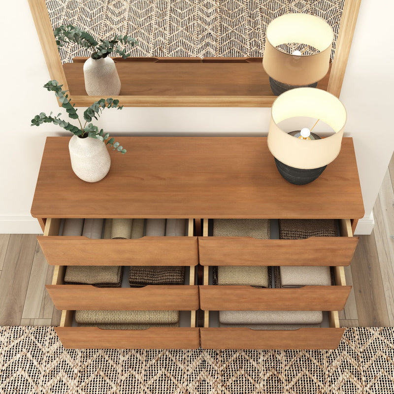 Wooden dresser with open drawers displaying folded clothes, adorned with a potted plant and table lamp, set against a geometric-patterned rug and mirror.