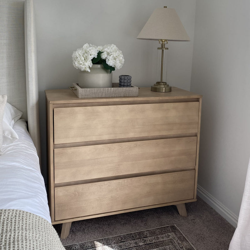 Modern light-wood three-drawer dresser in neutral bedroom with beige table lamp and white hydrangea bouquet in ceramic vase on decorative tray, minimalist Scandinavian décor