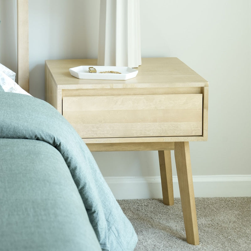Mid-century modern wooden nightstand with drawer next to a bed in a cozy bedroom setting.