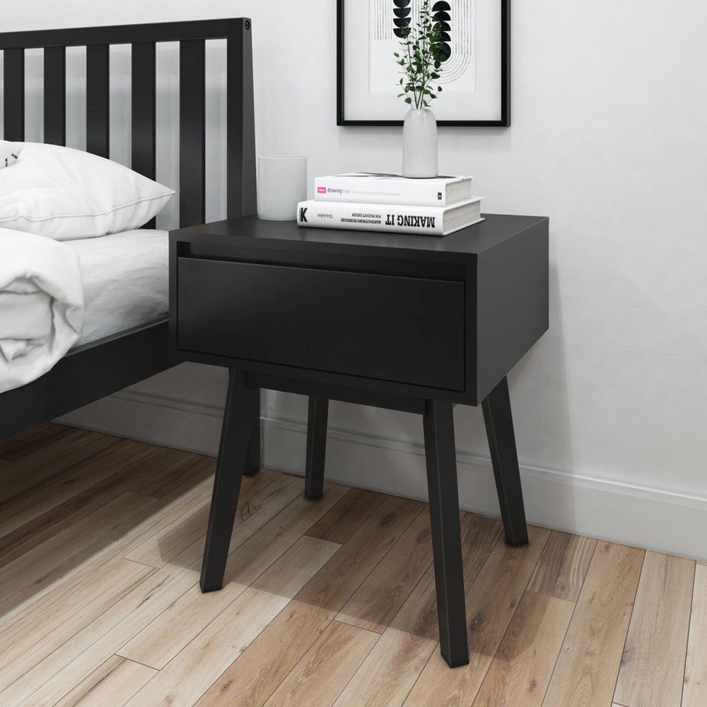 Black wooden bedside table with drawer, holding books and a vase, next to a black bed on hardwood floor in modern bedroom.
