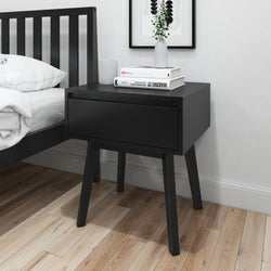 Black wooden bedside table with drawer, holding books and a vase, next to a black bed on hardwood floor in modern bedroom.