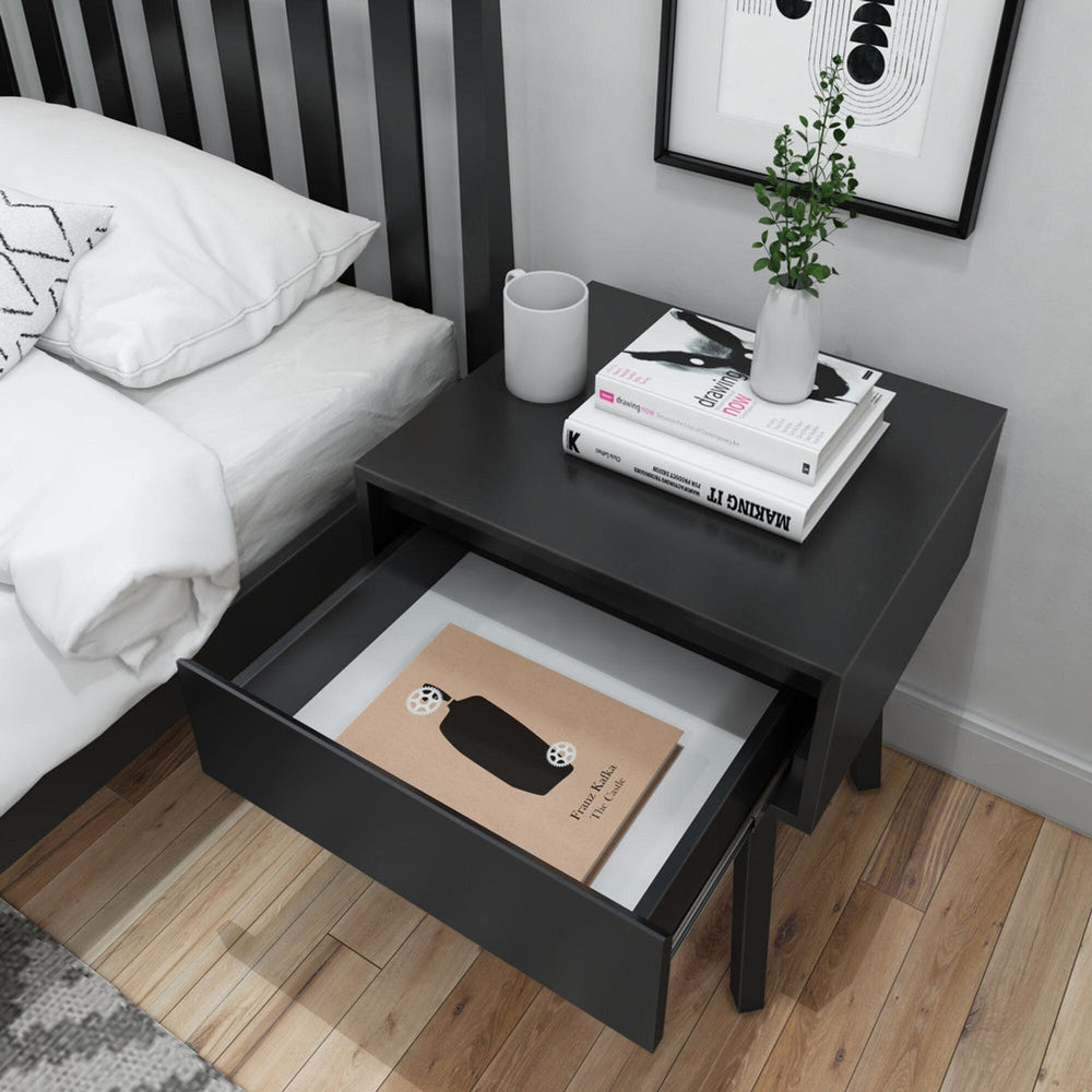 Modern black nightstand beside bed with books, plant, and open drawer displaying magazine in minimalist bedroom.