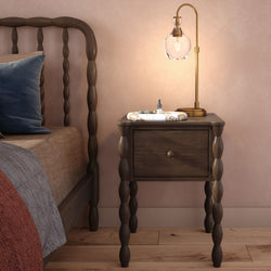 Cozy neutral bedroom nightstand with wooden spindle bed frame, dark wood bedside table, marble tray with skincare bottle, blue velvet pillow and modern brass table lamp against a soft pink wall