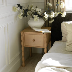 White hydrangea arrangement in textured ceramic vase on wooden bedside table with open book in a cozy neutral bedroom with beige curtains and natural light
