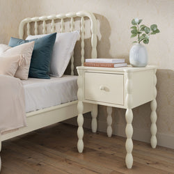 Cream spindle-leg bedside table with single drawer next to matching cream bed with decorative pillows, white vase with eucalyptus and stacked books in a neutral bedroom on hardwood floor