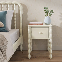 Cream spindle-leg bedside table with single drawer next to matching bed, vase with eucalyptus and stacked books on top, beige scallop wallpaper and hardwood floor