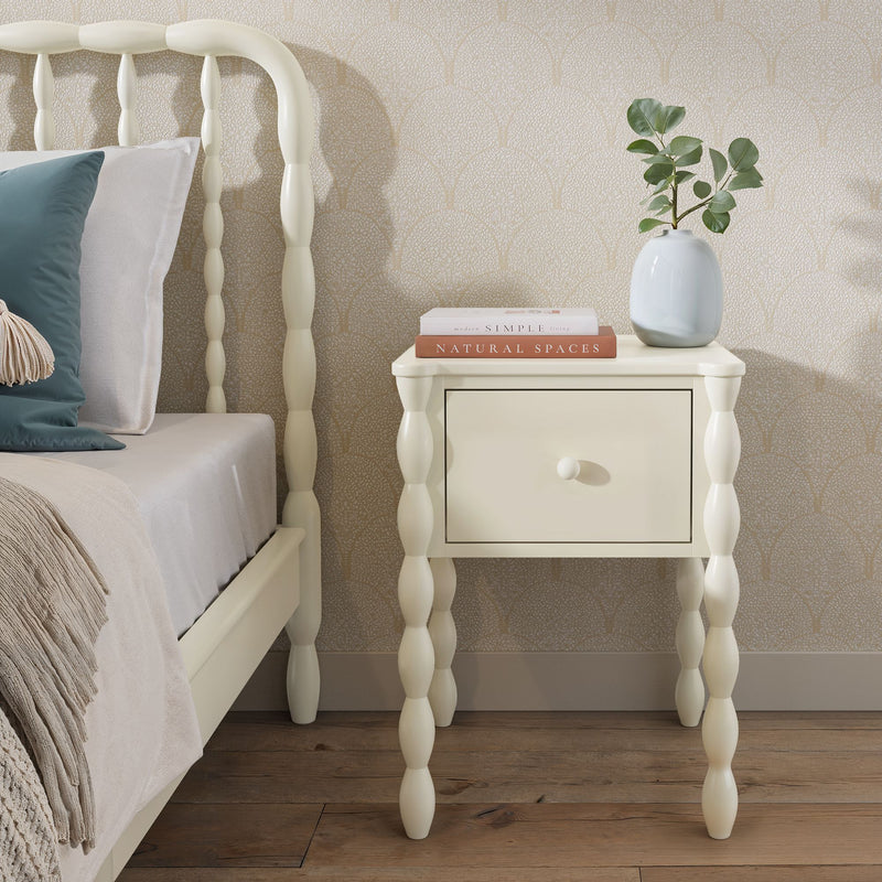Cream spindle-leg bedside table with single drawer next to matching bed, vase with eucalyptus and stacked books on top, beige scallop wallpaper and hardwood floor