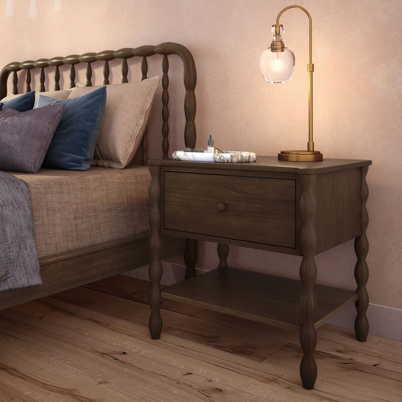 Rustic wooden bedside table with drawer and brass glass lamp beside spindle wood bed, textured neutral pillows and oak hardwood floor in pink-accent bedroom