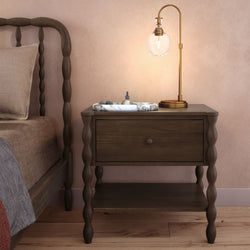Rustic dark wood bedside table with turned legs and drawer, brass glass globe bedside lamp, marble tray with dropper bottle on hardwood floor against a pink textured bedroom wall