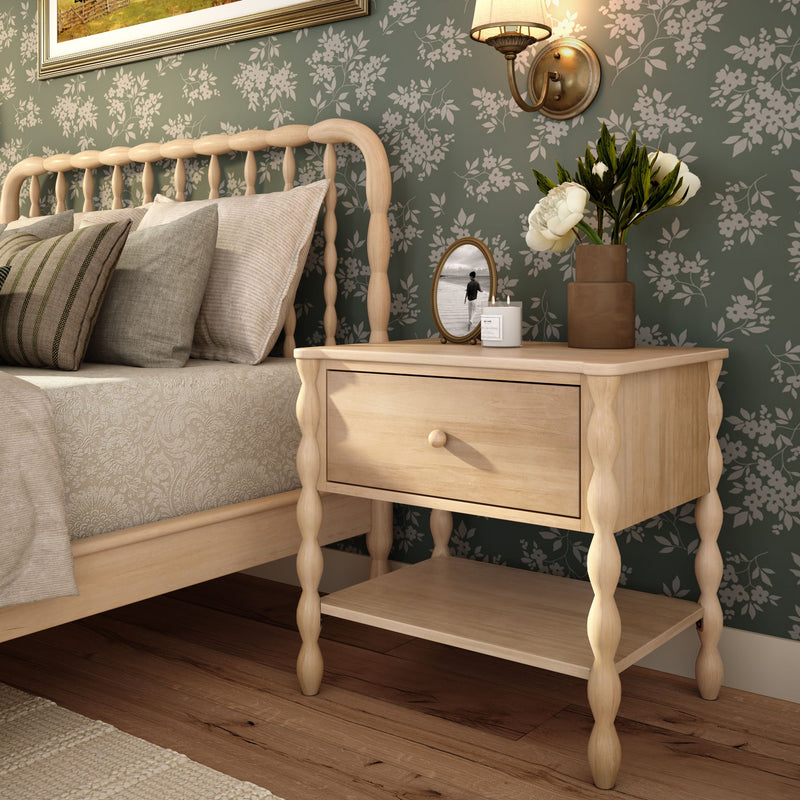 Cozy farmhouse bedroom with spindle wooden bed and light wood nightstand, green floral wallpaper, neutral bedding, white peony vase, candle and brass wall sconce on hardwood floor