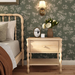 Cozy rustic bedroom with light wood spindle bed and bedside table against green floral wallpaper, wall sconce, vase of white peonies, candle and framed black-and-white photo