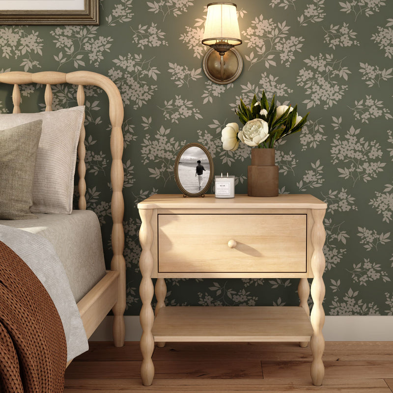 Cozy rustic bedroom with light wood spindle bed and bedside table against green floral wallpaper, wall sconce, vase of white peonies, candle and framed black-and-white photo