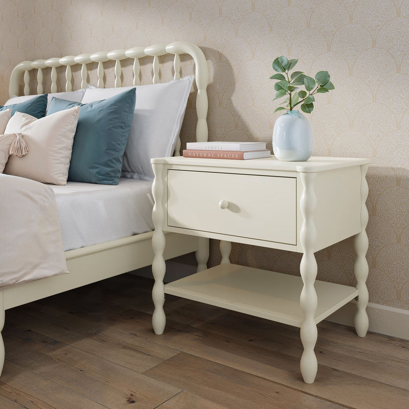 Cream spindle bedside table with drawer and lower shelf beside matching spindle bed in farmhouse bedroom with teal pillows, eucalyptus vase and wood flooring